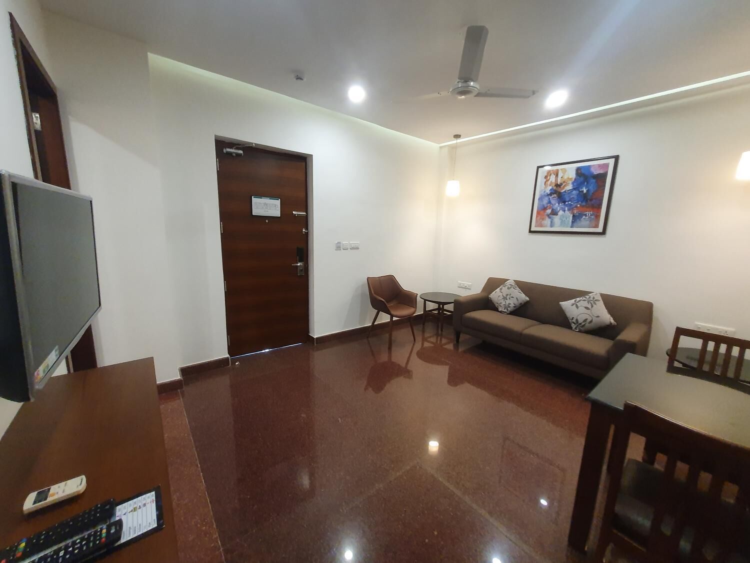 A modern living room with a flat-screen TV, a wooden door, a brown sofa, a small table, and a ceiling fan.