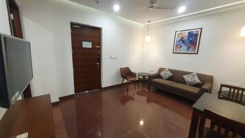 A modern living room with a flat-screen TV, a wooden door, a brown sofa, a small table, and a ceiling fan.