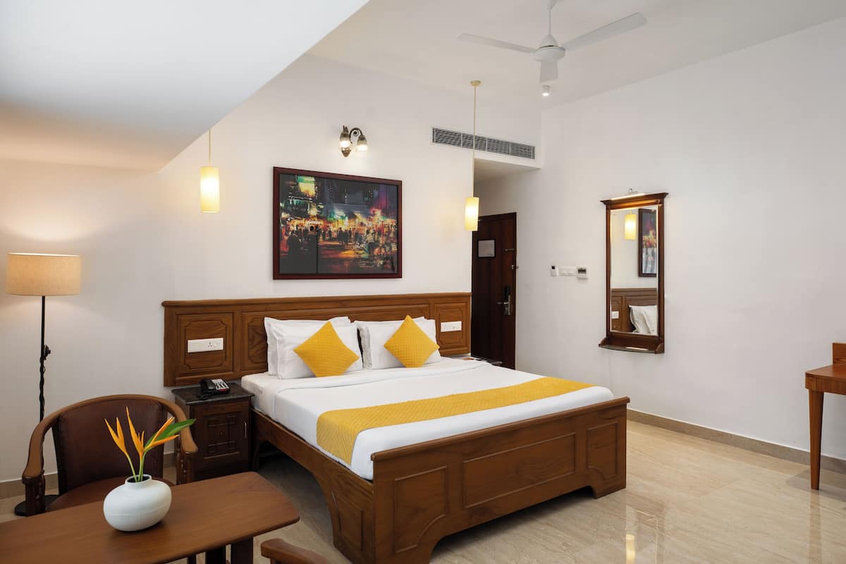 Una habitación de hotel con cama de madera, una mesita de noche, un escritorio, una silla, una lámpara, un cuadro, un espejo y un ventilador de techo.