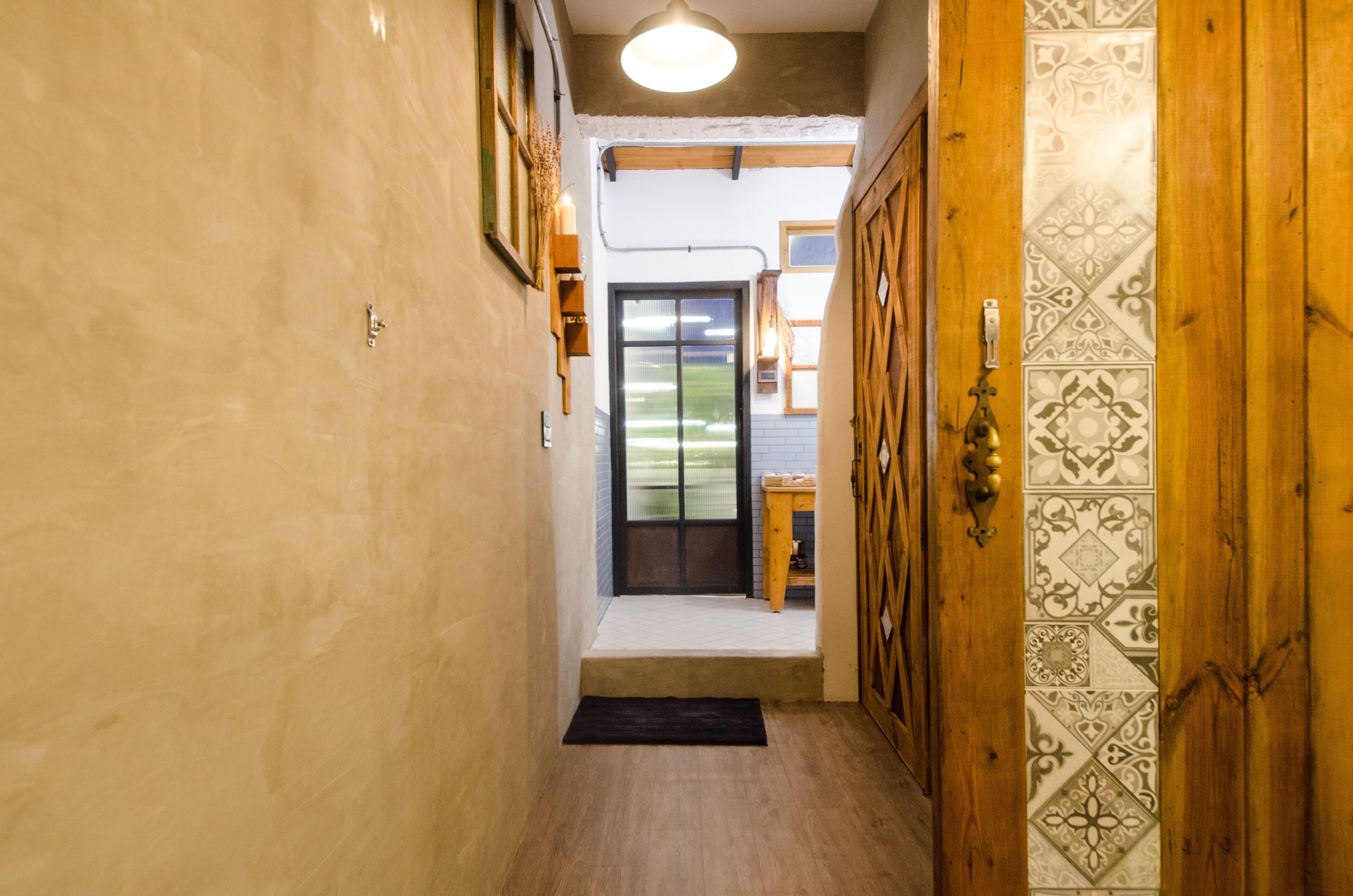 Un pasillo estrecho con una puerta de madera, un felpudo negro en la entrada y vista a un jardín por una puerta de vidrio.