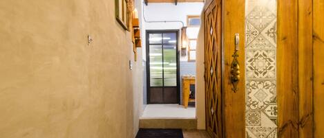 Un pasillo estrecho con una puerta de madera, un felpudo negro en la entrada y vista a un jardín por una puerta de vidrio.
