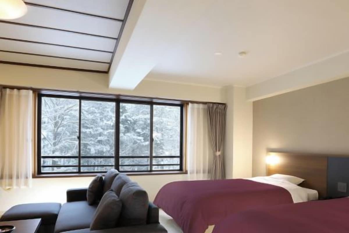 Une chambre d’hôtel avec un canapé, deux lits, un bureau et une grande fenêtre donnant sur la forêt.