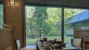 Perinteinen japanilaistyylinen huone, jossa on tatamimatto, matala pöytä teetarvikkeineen ja näkymä vehreään puutarhaan.