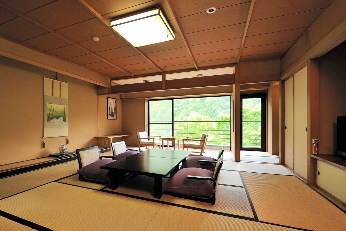 Une pièce de style japonais traditionnel, avec un sol recouvert de tatamis, une table basse et des portes coulissantes donnant sur un balcon offrant une vue sur la verdure.