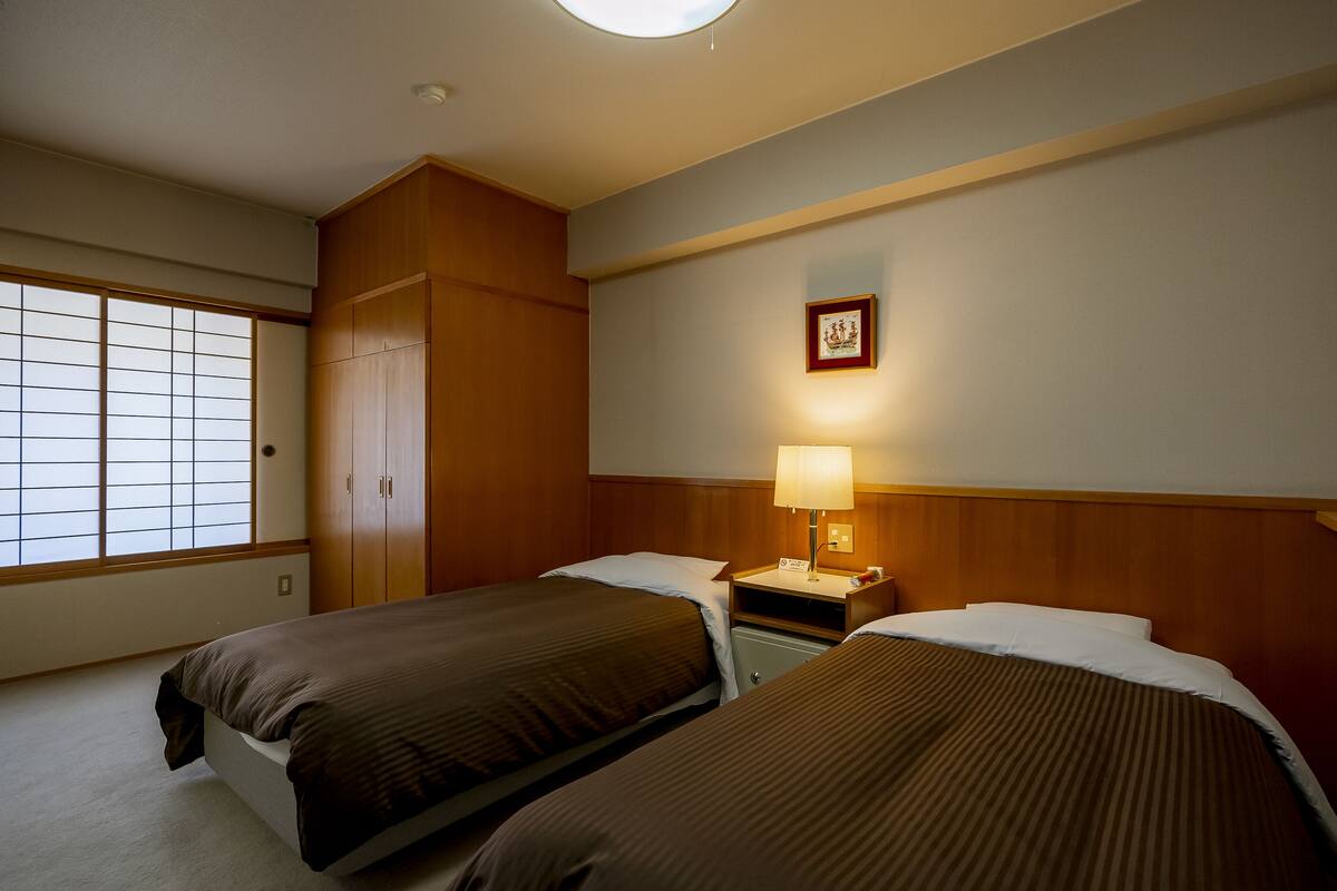 Habitación de hotel con dos camas, un armario de madera, una mesita de noche con una lámpara y un cuadro en la pared.