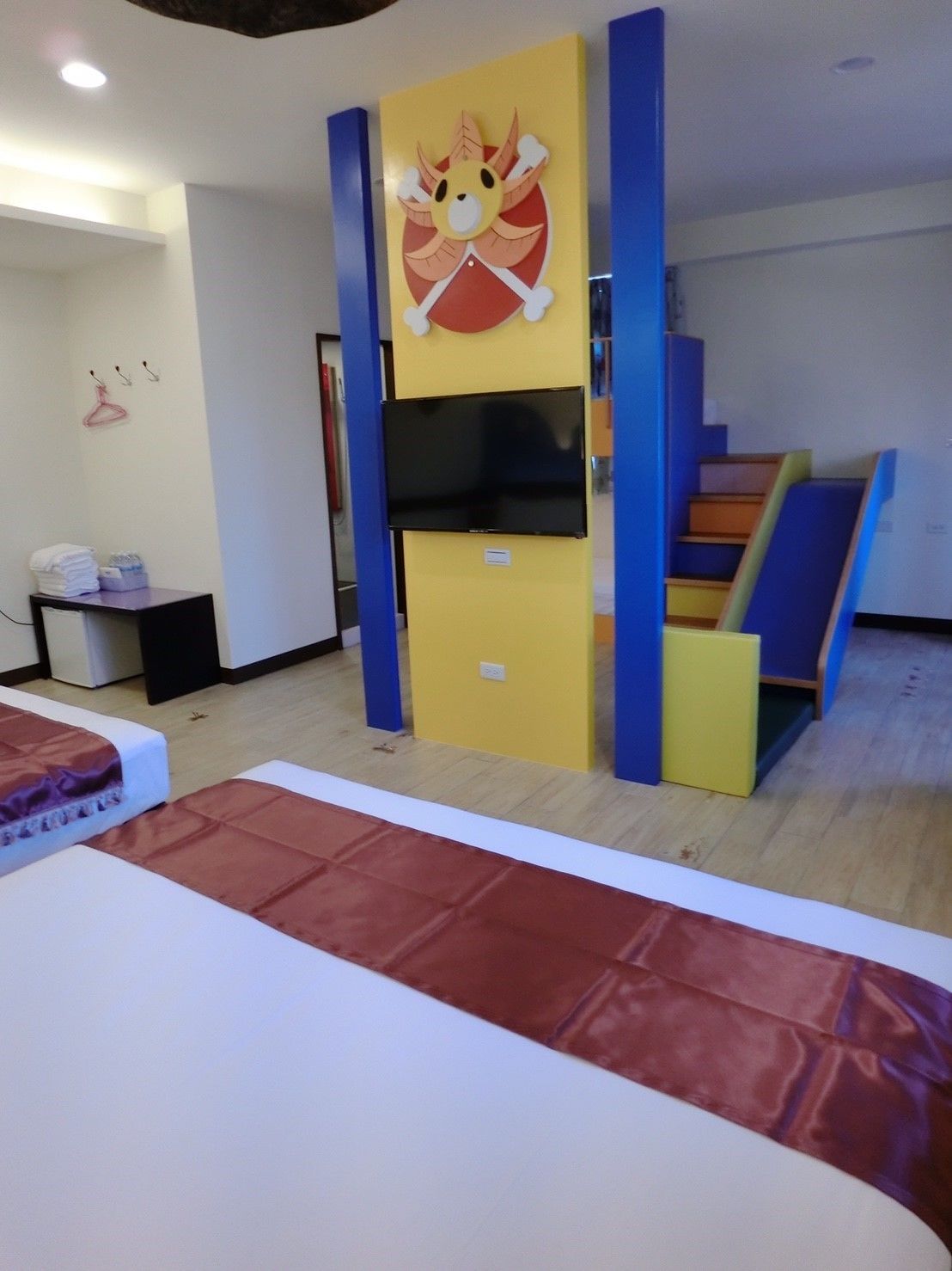 Una habitación con una cama, un televisor en un mueble amarillo y azul, una mesita con objetos y una escalera.