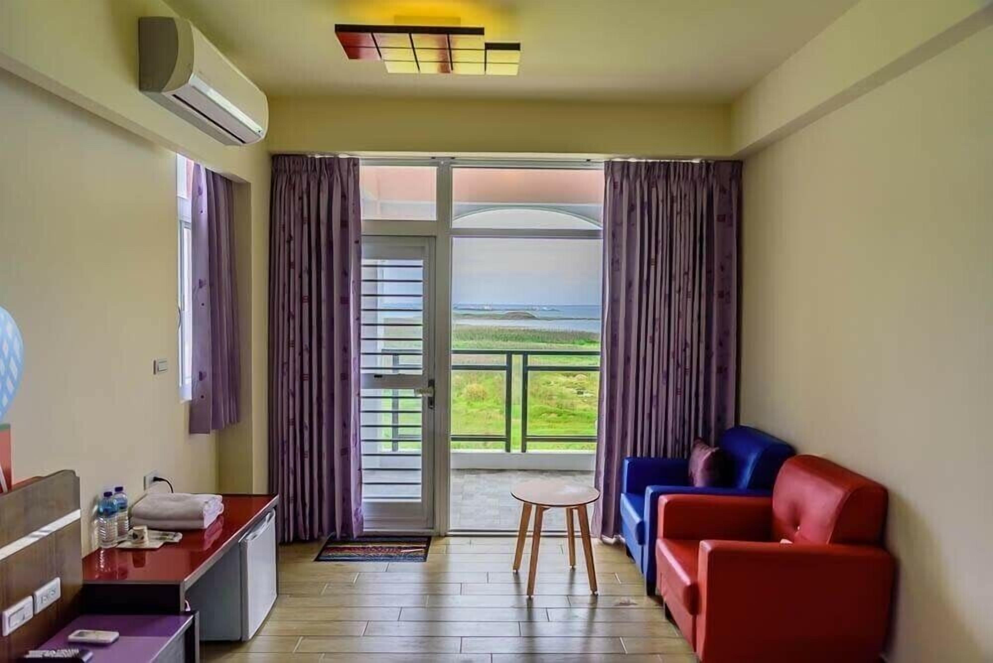 Una habitación con escritorio, sofá rojo y azul, una mesita y vistas al exterior.
