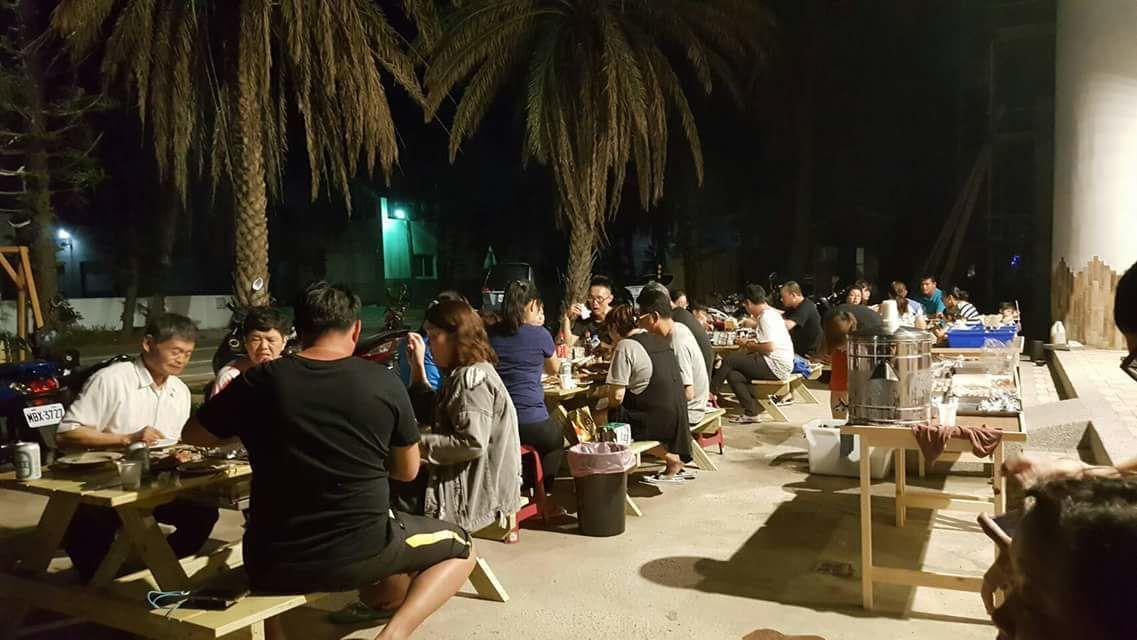 An outdoor dining area with tables and chairs, palm trees, and a stage in the background.