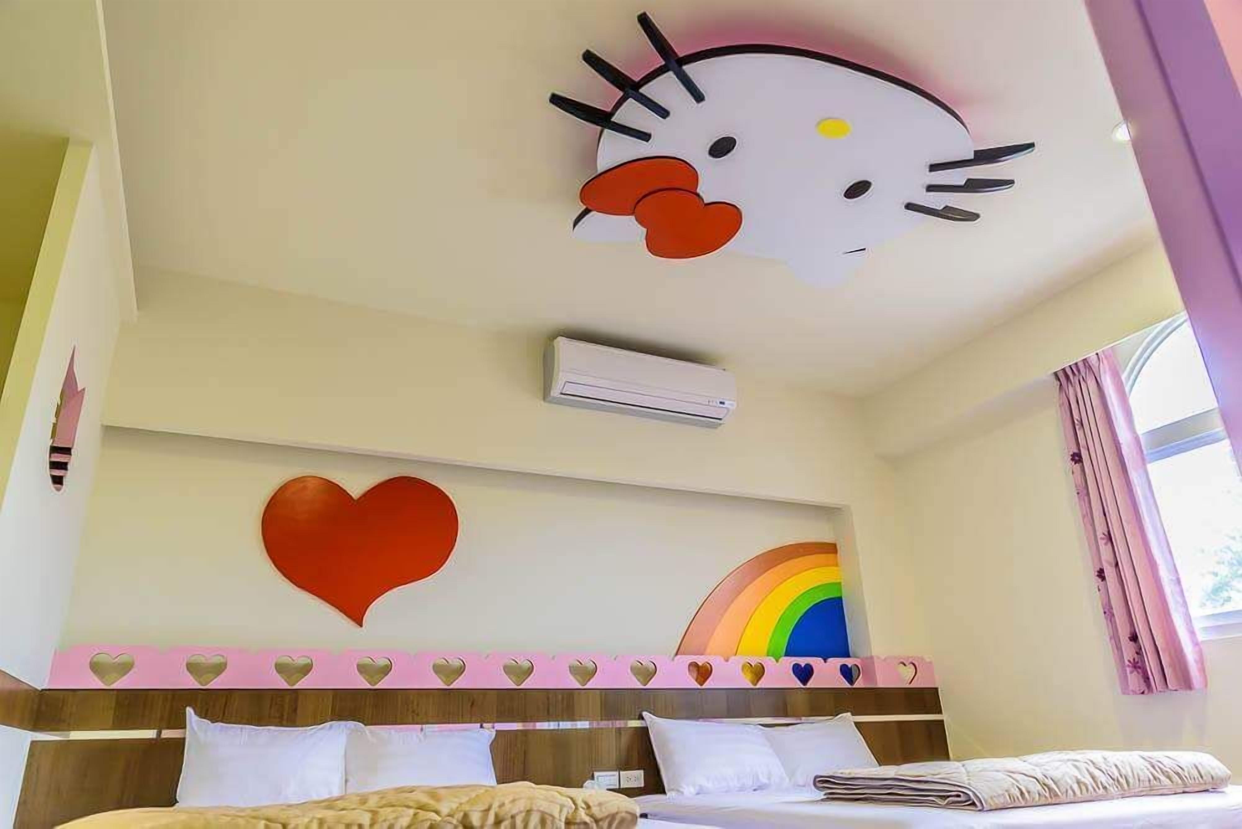 A hotel room with a bed, a wall-mounted air conditioner, a large Hello Kitty decoration, a heart-shaped decoration, and a rainbow design.