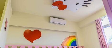 A hotel room with a bed, a wall-mounted air conditioner, a large Hello Kitty decoration, a heart-shaped decoration, and a rainbow design.