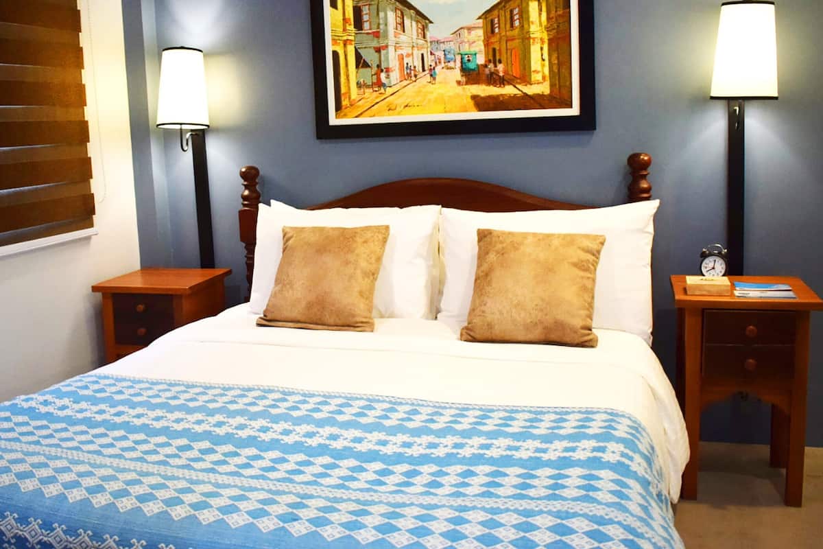 A hotel room with a large bed, two nightstands, a painting on the wall, and a window with blinds.
