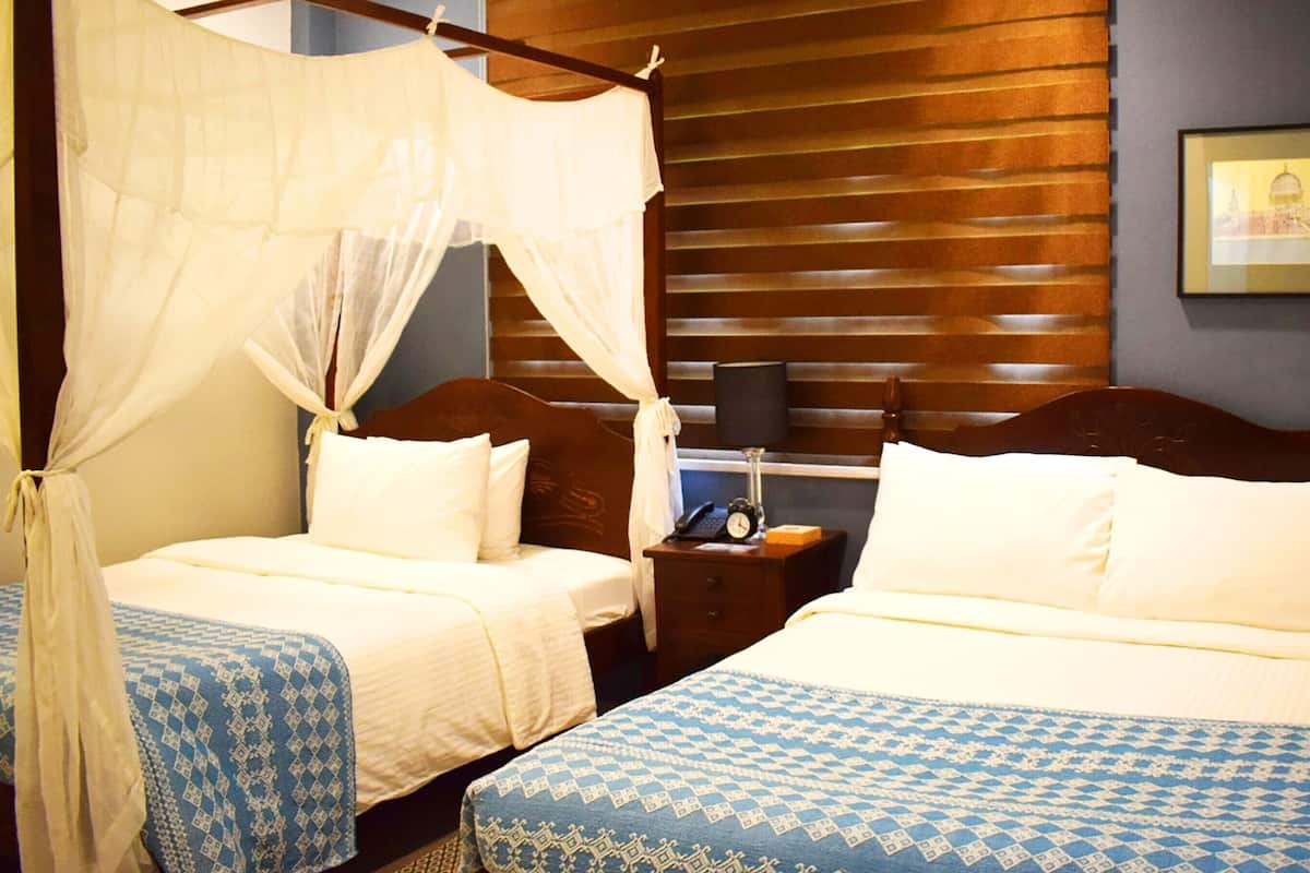 A hotel room with two beds, one with a canopy and the other with a headboard. There is a nightstand with a lamp and a clock.