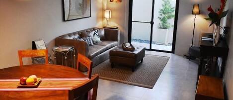 A living room with a sofa, coffee table, and dining table with fruit. There is a sliding glass door leading to an outdoor area.