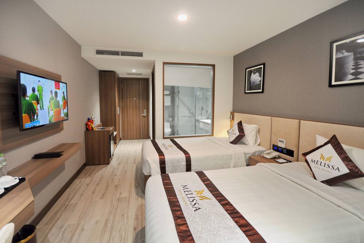 A hotel room with two beds, a TV, a desk, and a bathroom visible through a glass door.