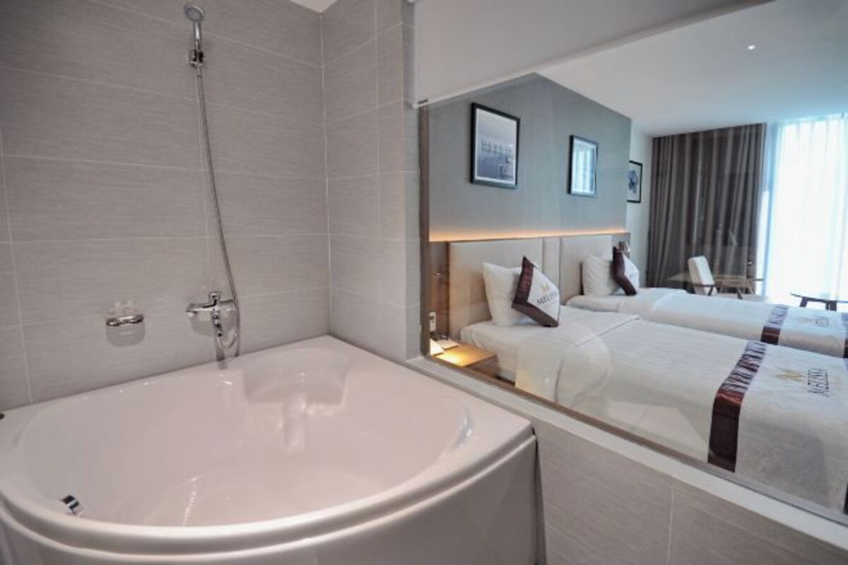 A modern hotel room with a large bathtub, two beds, a nightstand, and a large window with curtains.