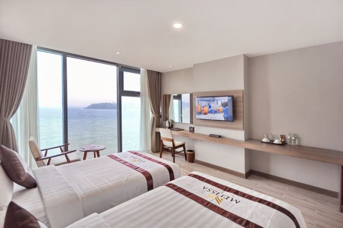 Habitación de hotel con dos camas, televisión, escritorio y vista al mar.