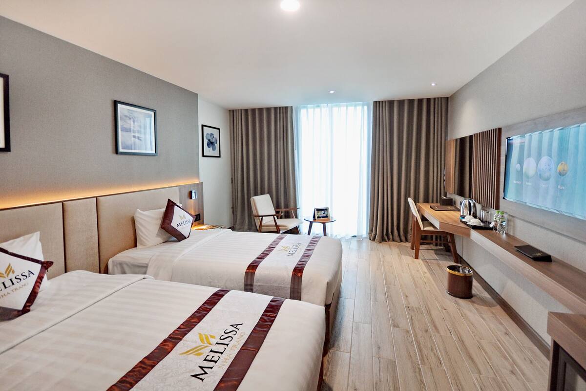 A hotel room with two beds labeled 'Melissa', a desk, a chair, a TV, and a window with curtains.