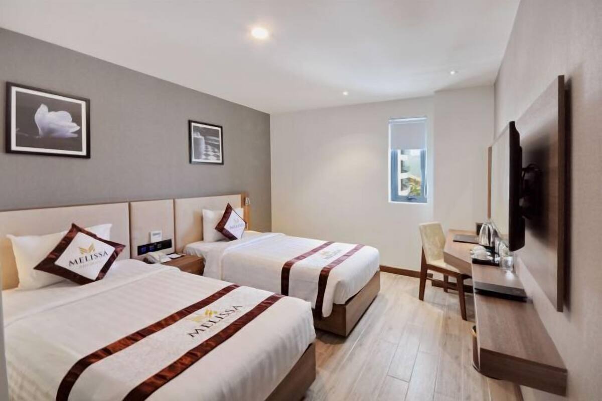 A hotel room with two beds, a desk, a chair, a TV, and framed pictures on the wall.