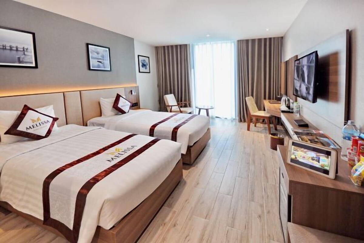 A hotel room with two beds, a TV, a desk with a computer, and a window with curtains.