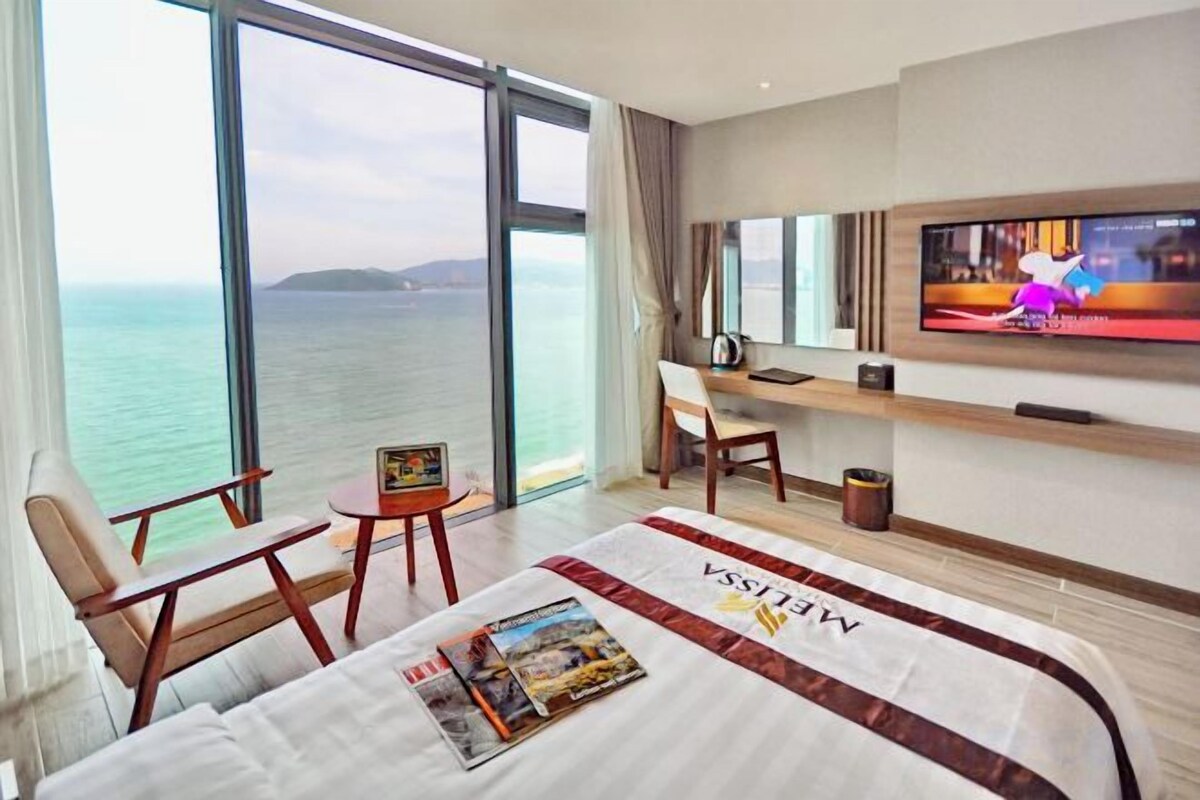 A hotel room with a bed, a TV, a desk, and a view of the sea.