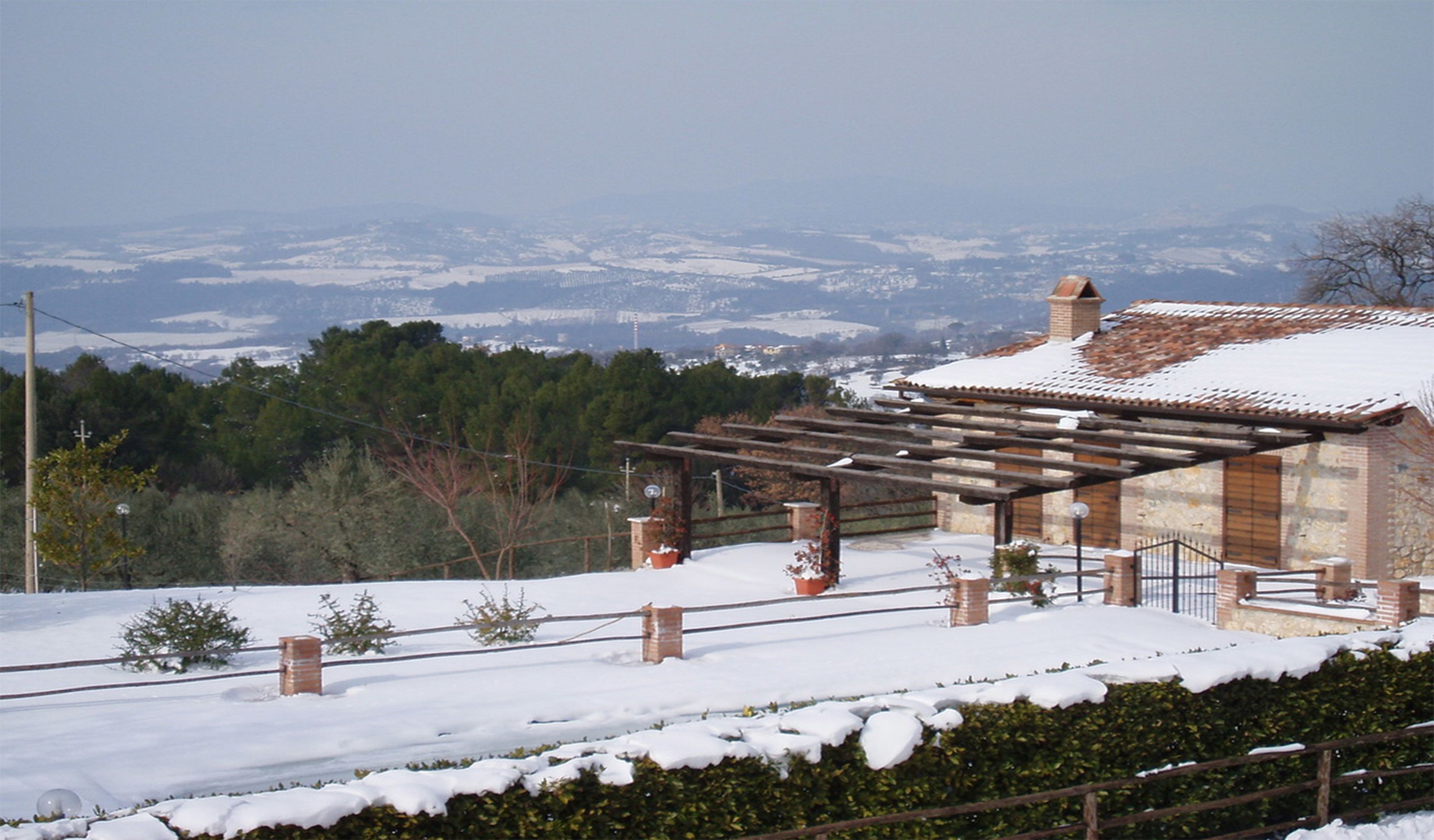Un edificio con pérgola en una ladera nevada, rodeado de árboles y una vista distante de un valle.