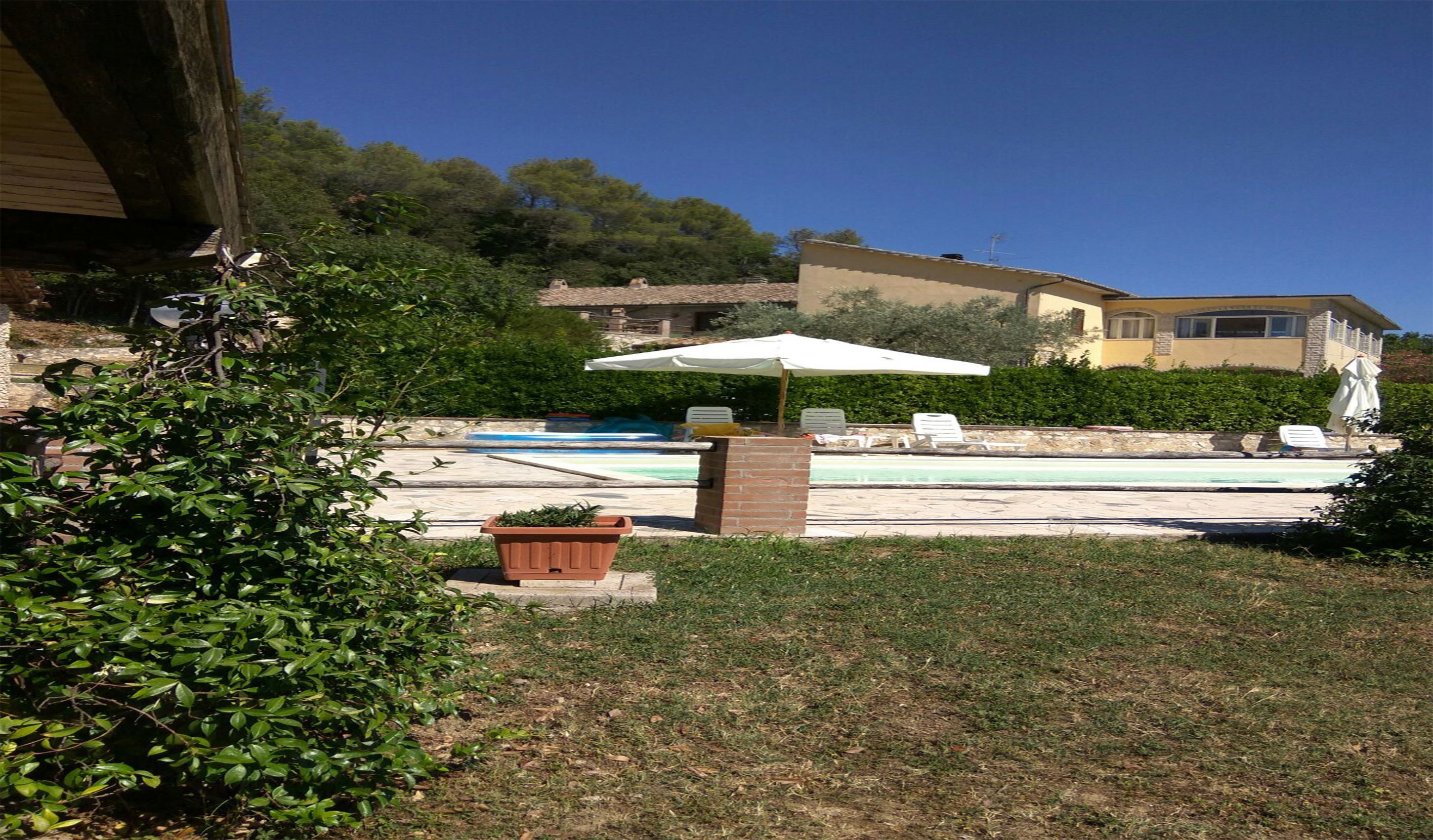 A pool area with lounge chairs and umbrellas, surrounded by greenery and a building in the background.