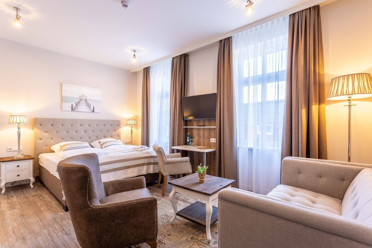 A hotel room with a bed, two armchairs, a small table, a TV, and a window with curtains.