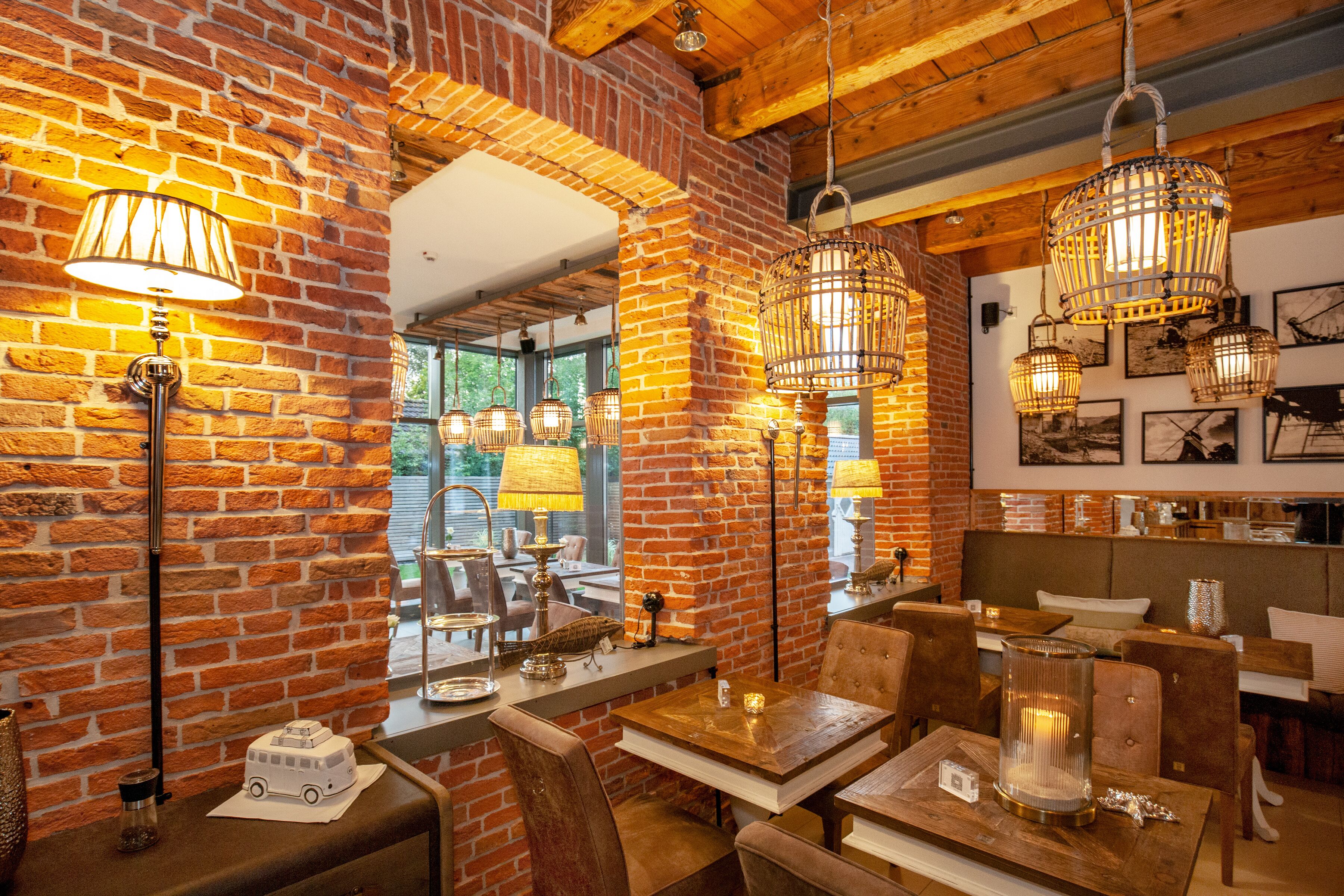 Een restaurant met bakstenen muren, houten balken en hangende lampen. Tafels staan klaar met stoelen, en er hangen afbeeldingen in lijsten aan de muren.