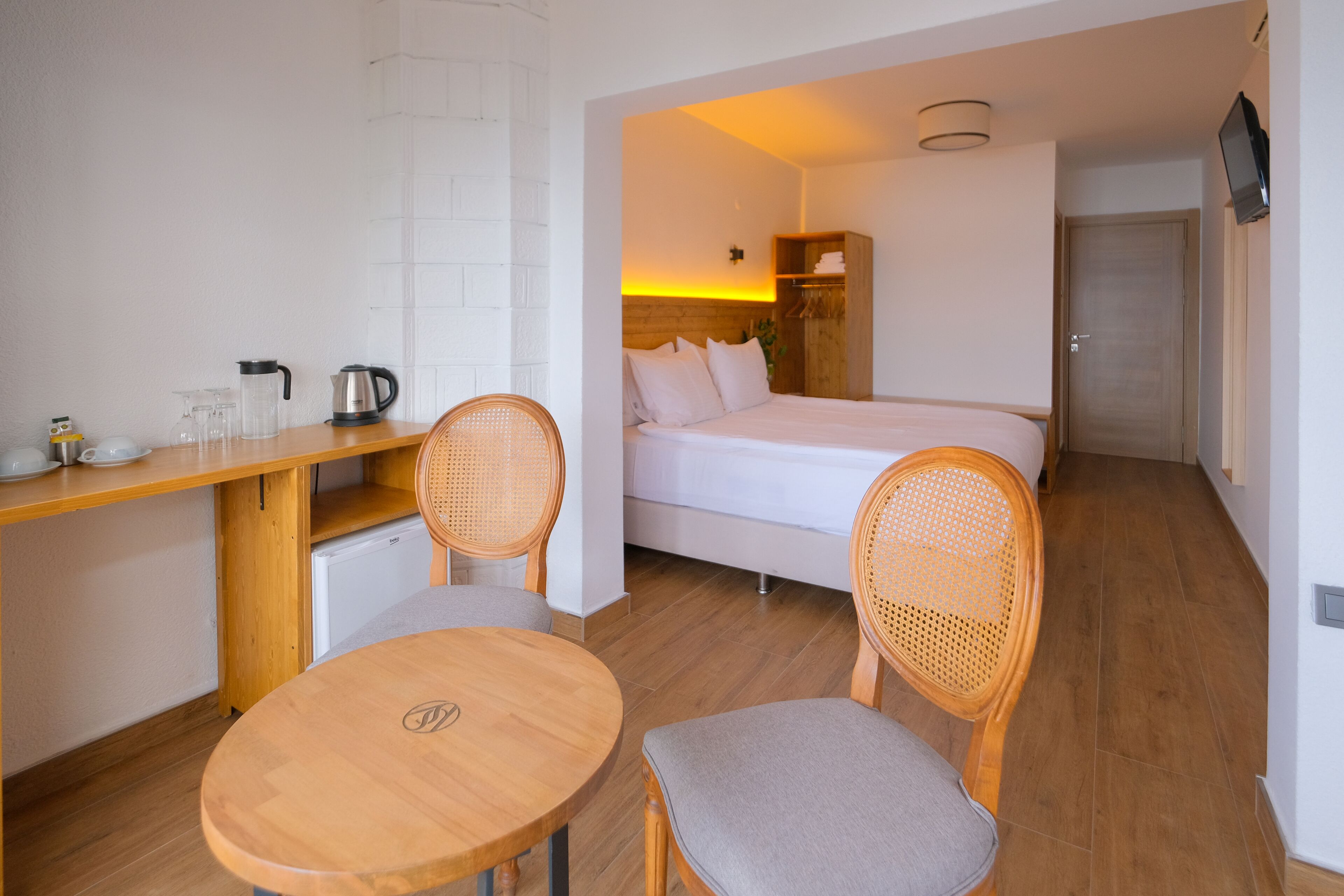 Una habitación de hotel compacta con una cama, una kitchenette pequeña, una zona de comedor con mesa y sillas, y un televisor montado en la pared.