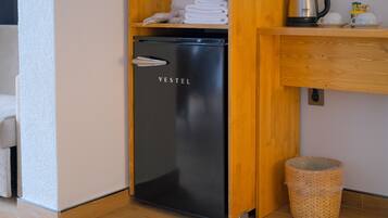 A compact refrigerator with a handle, a wooden shelf above it holding folded towels and a kettle, and a wicker basket on the floor.