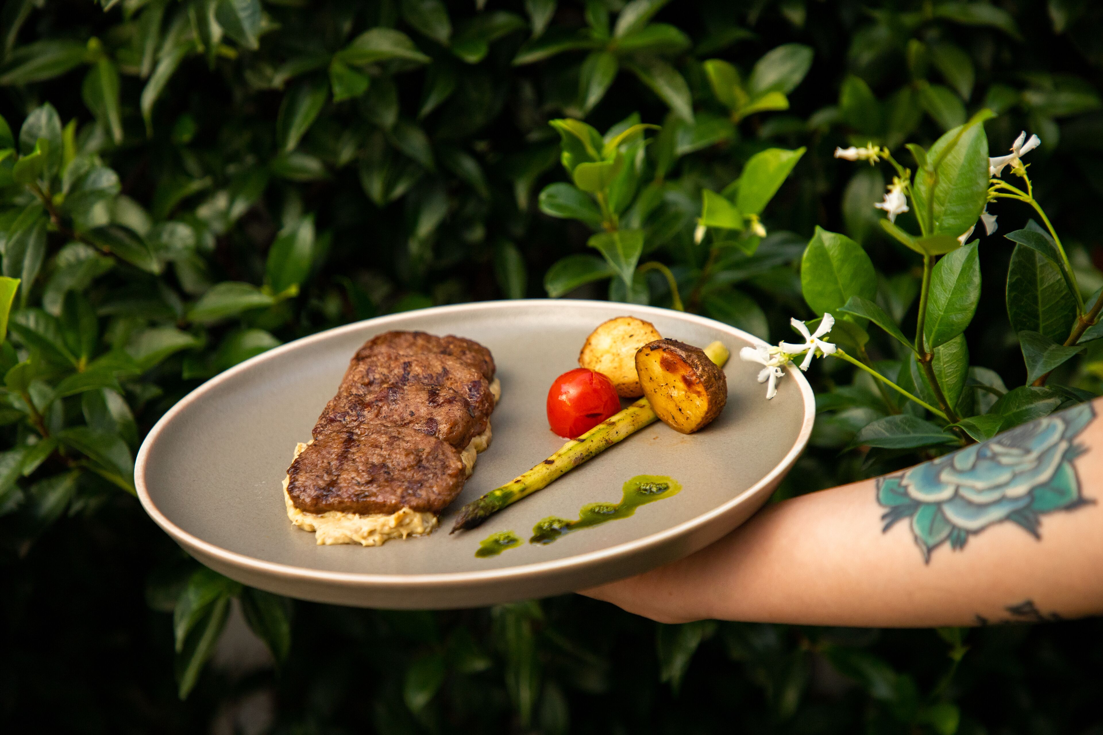 Un plato con un bistec, espárragos y tomate, sostenido por una persona con un tatuaje.