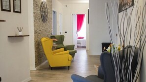 Una sala de estar moderna con piso de madera, un sillón amarillo, un sillón verde y un sillón azul. Se observa una pieza decorativa en la pared y un estante pequeño con objetos a la izquierda.