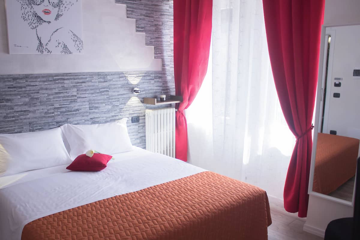 A hotel room with a bed, red curtains, a white wall with a textured design, and a radiator.