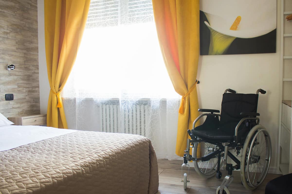 A bedroom with a bed, a wheelchair, and a window with yellow curtains.