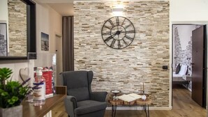 Una sala de estar moderna con una pared de piedra, un reloj grande, un sillón gris y una pequeña mesa de madera con velas.
