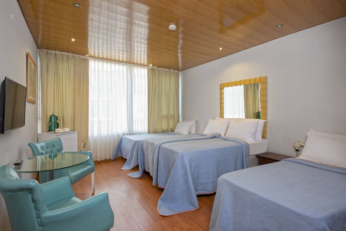 A hotel room with two single beds, a glass table with chairs, a small kitchenette, and a large window with curtains.