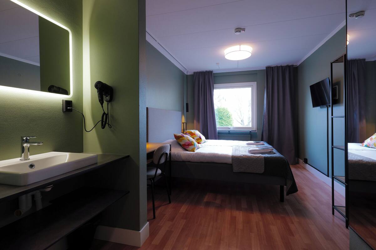Una habitación de hotel con un espejo grande, una cama con almohadas coloridas, una mesita de noche con una lámpara y una ventana con cortinas.