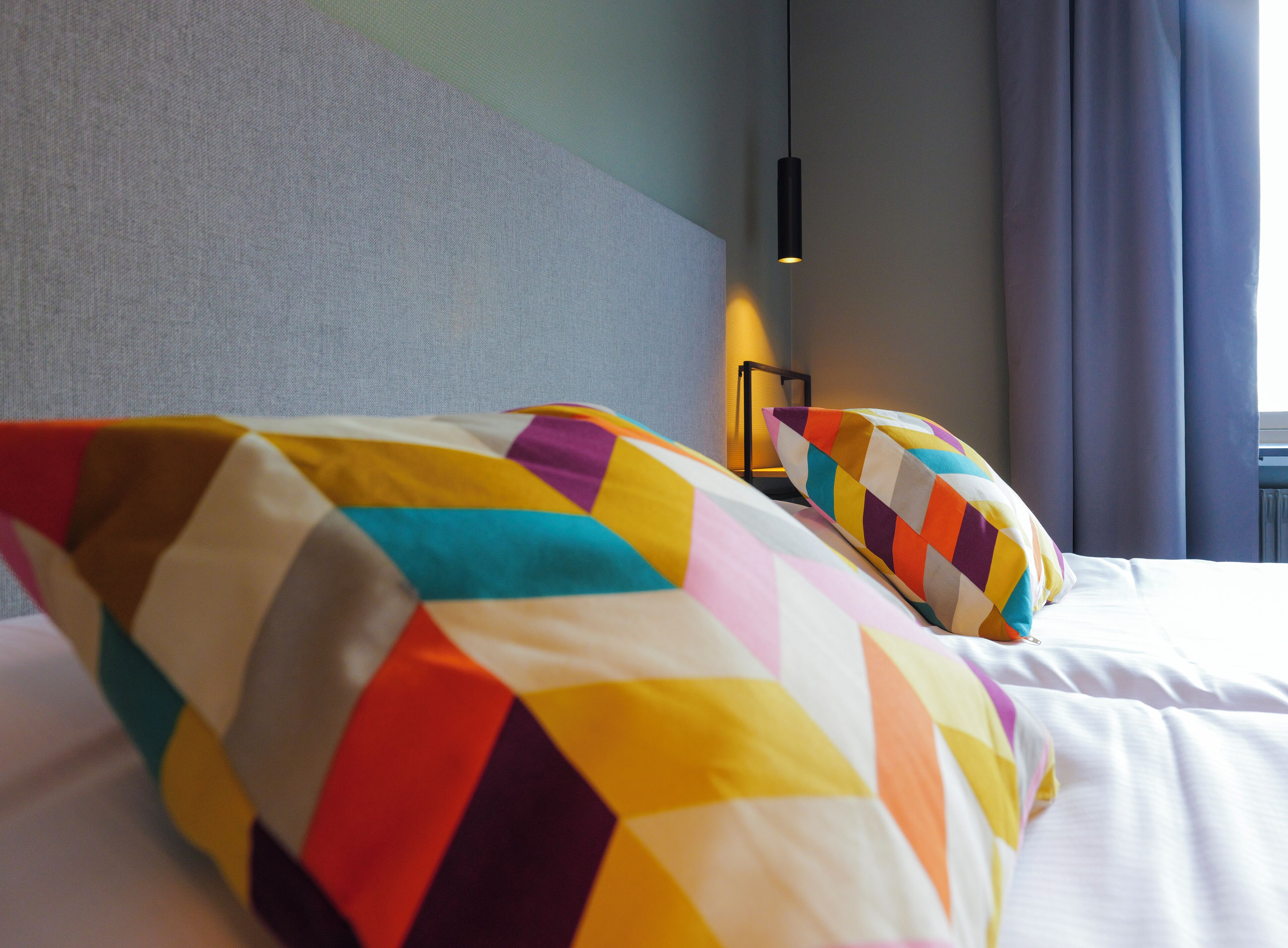 Una habitación de hotel con una cama bien hecha, almohadas coloridas, un cabecero y una lámpara de noche.