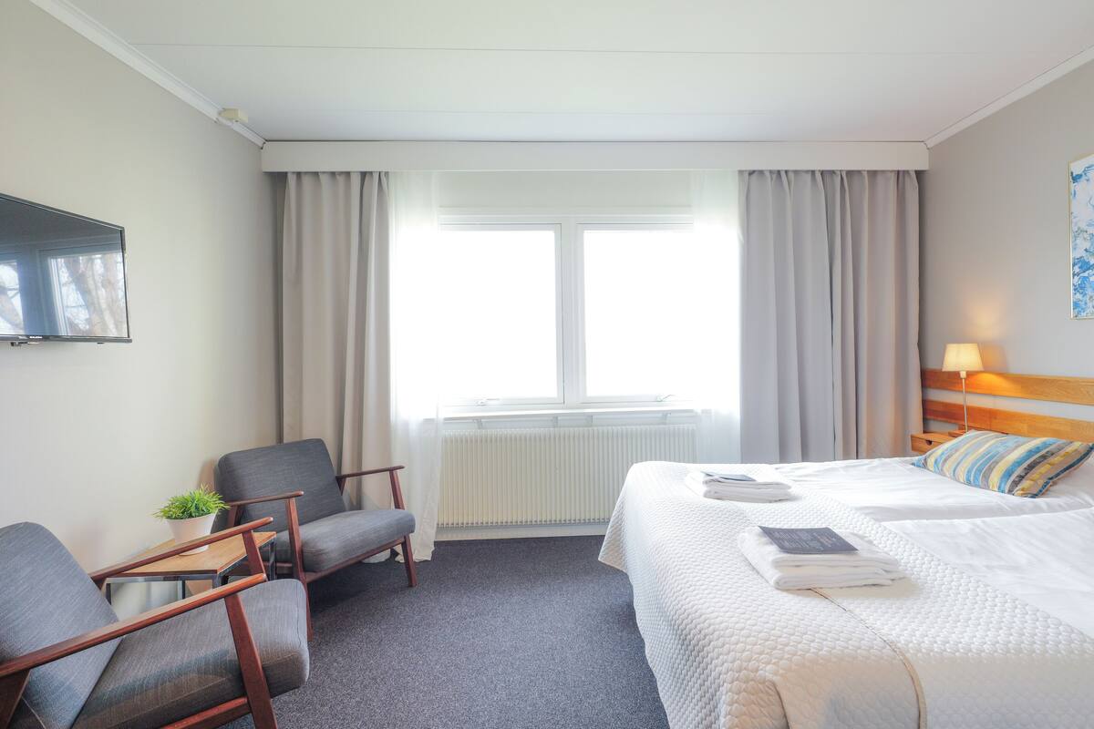 Habitación de hotel con una cama, una silla, una mesita con una planta, un radiador y una ventana con cortinas.