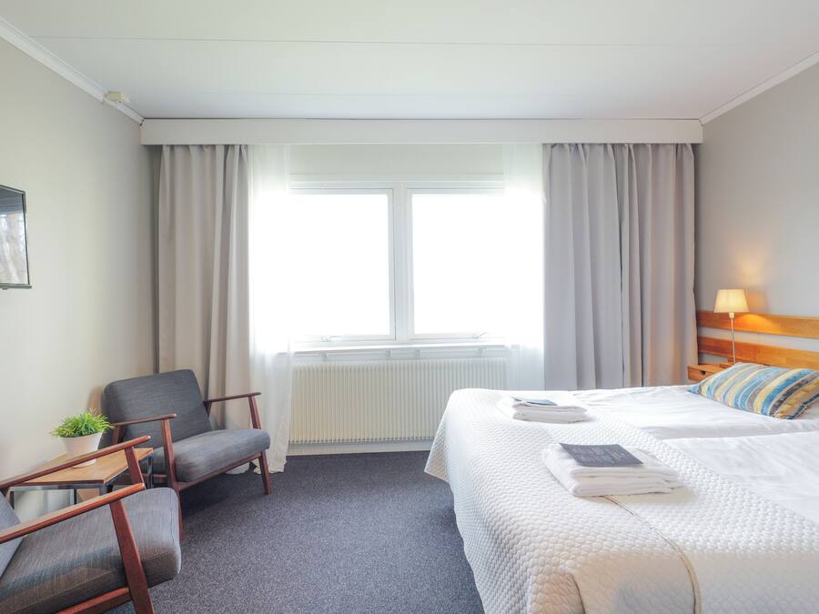 A hotel room with a bed, a chair, a small table with a plant, a radiator, and a window with curtains.