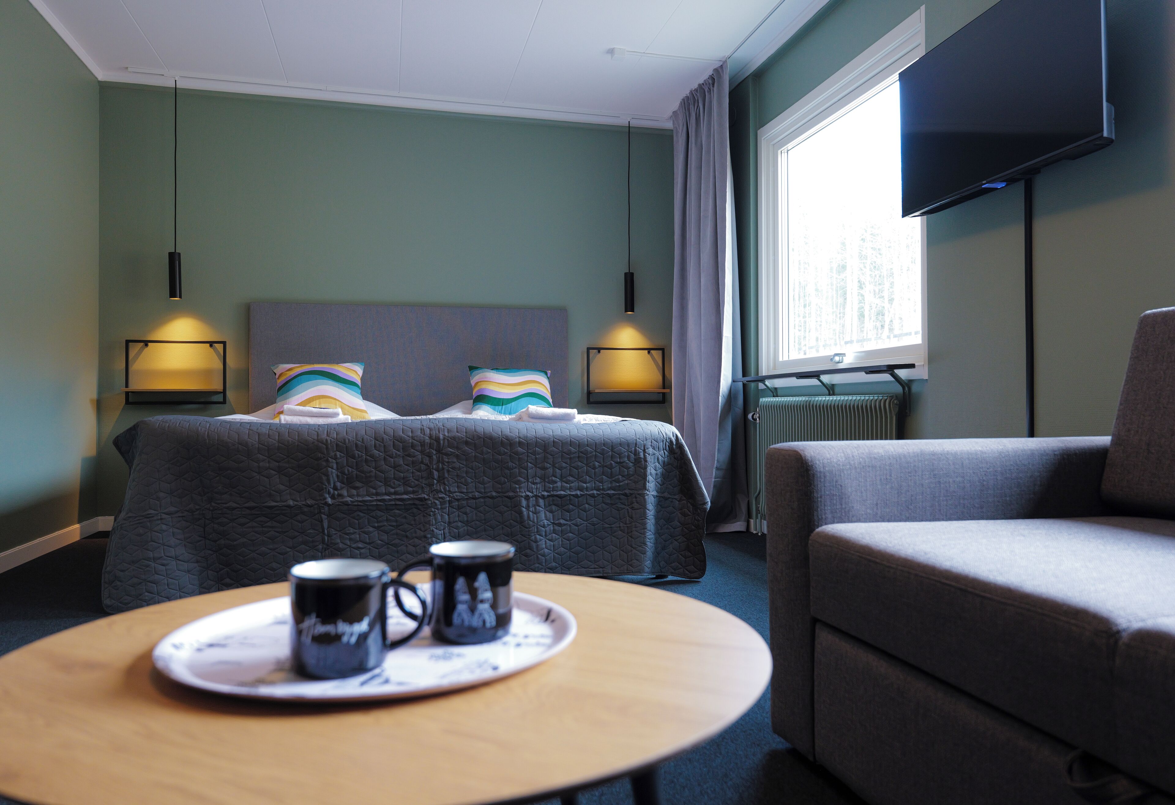 A hotel room with a bed, a sofa, a table with a tray and mug, and a TV mounted on the wall.