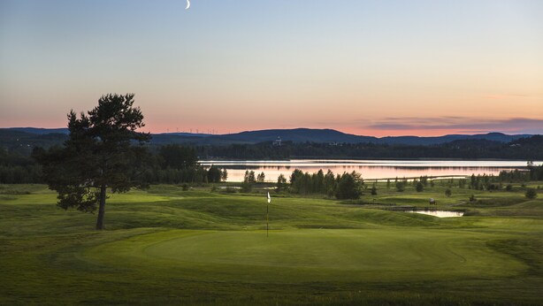 Ein Golfplatz mit Fahnenstange, einem Gewässer und Bergen in der Ferne.