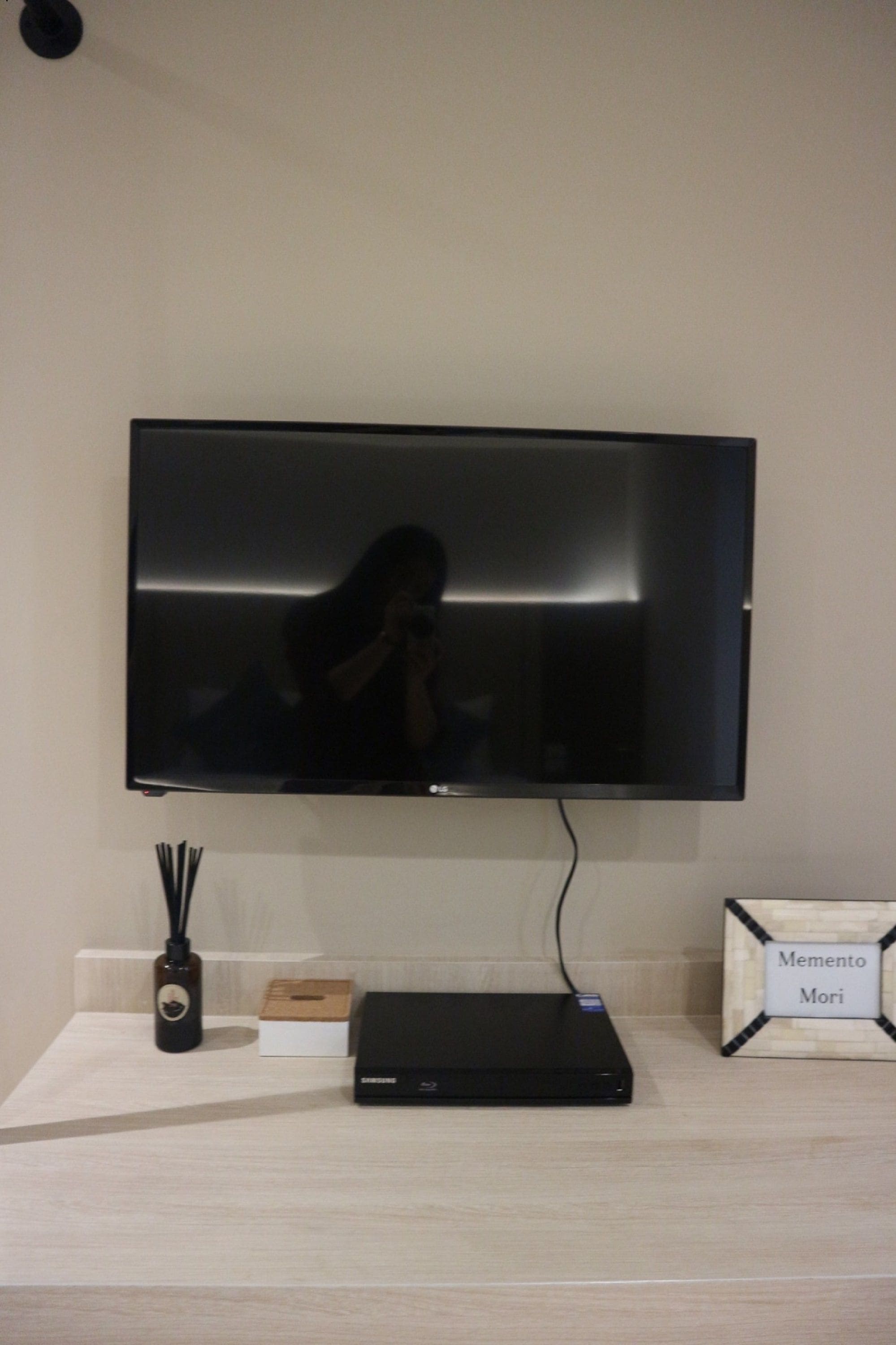 A flat-screen TV mounted on a wall, a black electronic device on a wooden table, and a small wooden box with the name "Memento Mori".