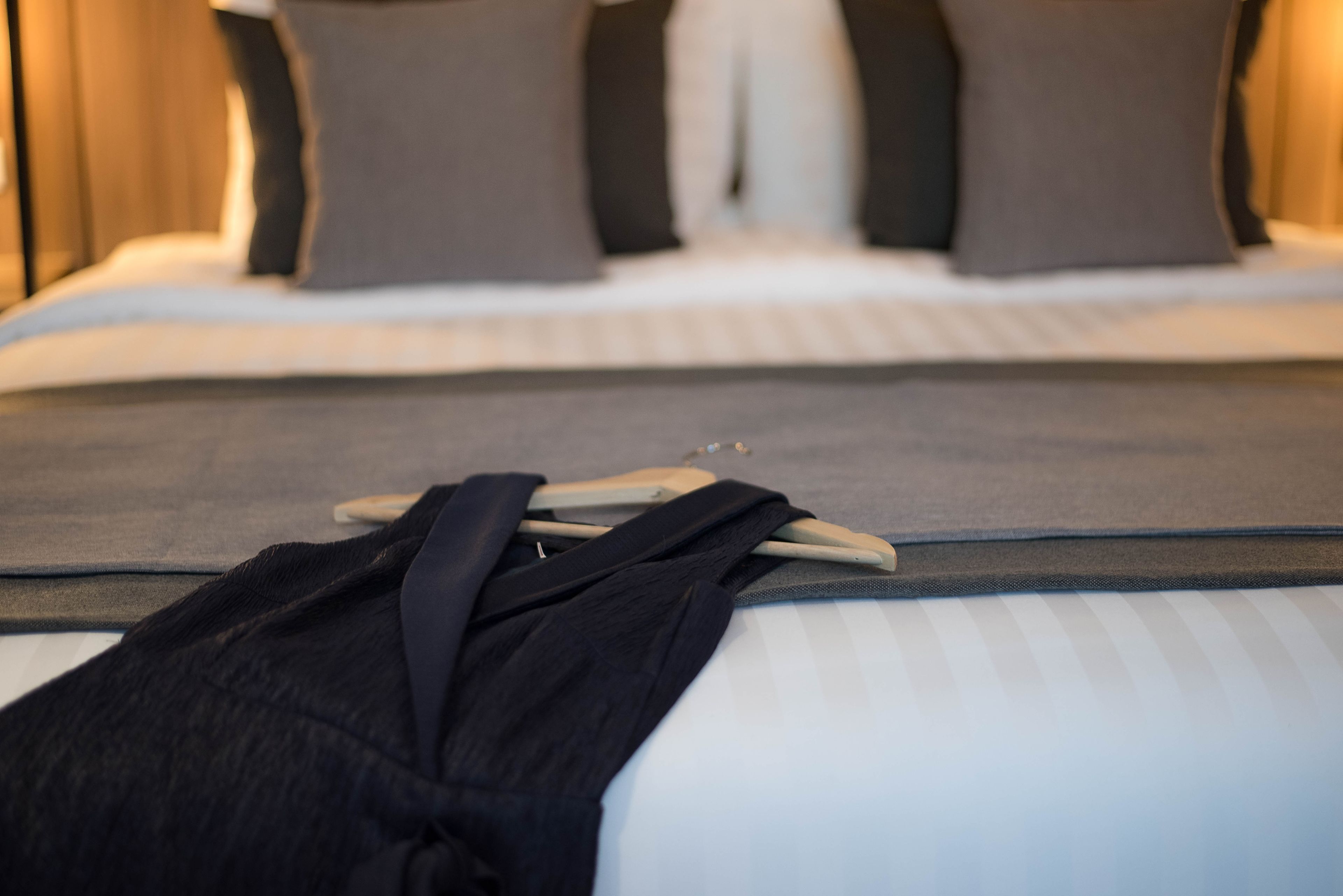 Een netjes opgemaakt bed met een grijze spreideken, een zwarte badjas over een kapstok en een houten hoofdbord.