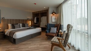 Una habitación de hotel moderna con una cama grande, una zona de estar con una silla y reposapiés, un armario y un ventanal con cortinas traslúcidas.
