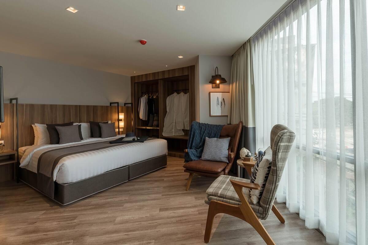 A modern hotel room with a large bed, a seating area with a chair and ottoman, a wardrobe, and a large window with sheer curtains.
