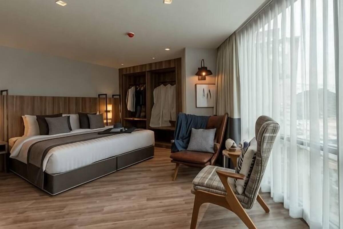 A modern hotel room with a large bed, a seating area with chairs, a wardrobe, and a view through large windows.