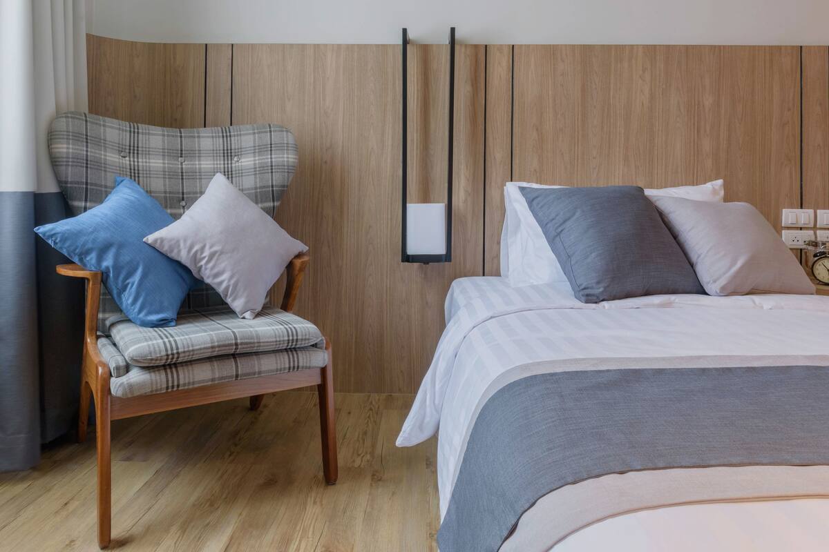 A modern bedroom with a wooden headboard, a bed with grey bedding, a chair with plaid cushions, and a bedside table.