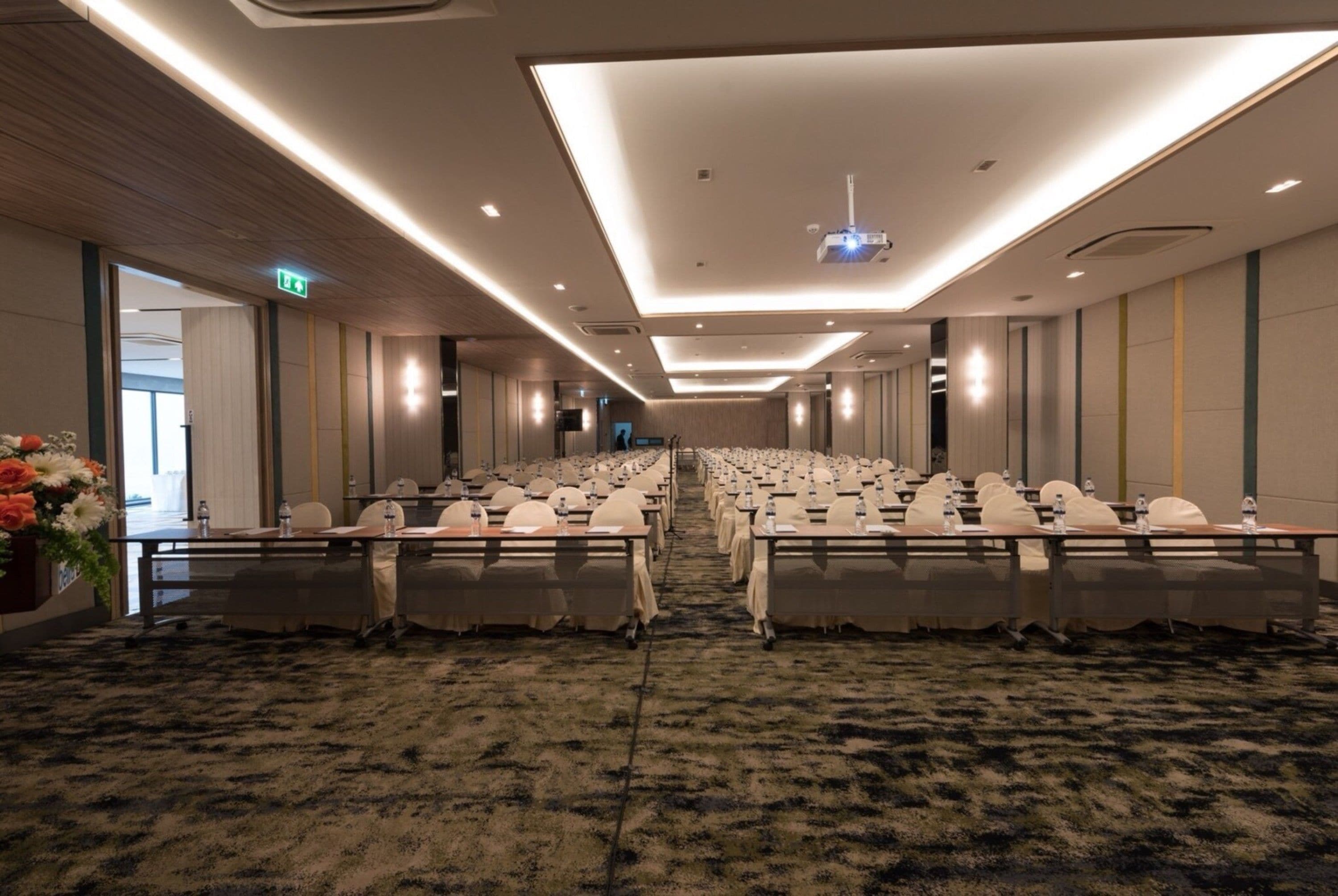 A conference room with rows of tables and chairs, a carpeted floor, and a ceiling with recessed lighting and a projector.