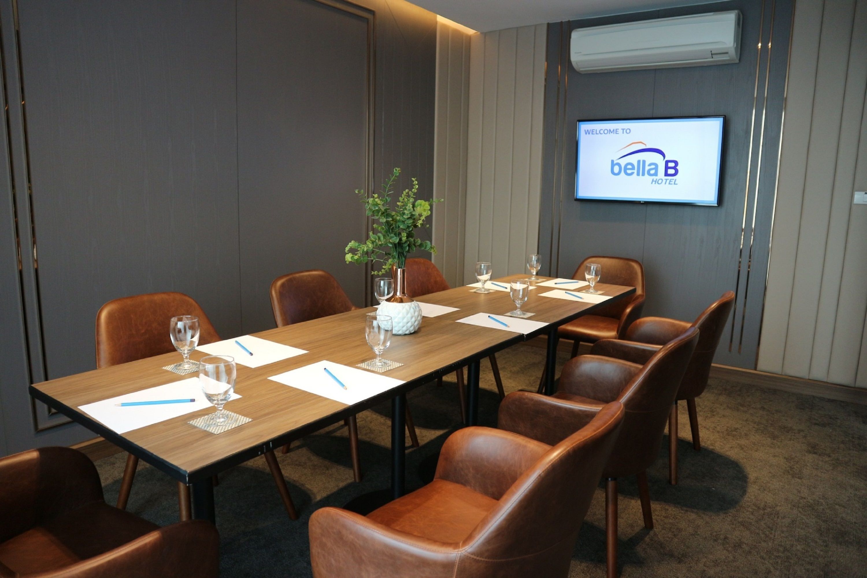 Un salón de conferencias con una larga mesa, sillas de cuero marrón y una pantalla que muestra “Welcome to Bella B Hotel”.